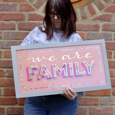 Katrin from Chic Sign Studio holding a reverse glass gilded sign artwork featuring the words We are Family in 23ct gold leaf on a pale pink background, infilled with dried flowers, abalone shell, glitter and finished in a real wood grey frame