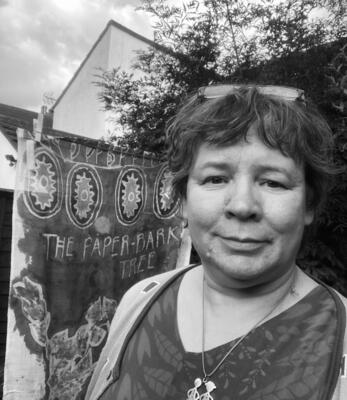 Black and white photo of textile artist Jane Charles, stood by a piece of batik fabric and lilac tree. 
