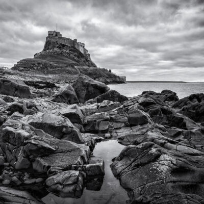 Stuart Feurtado: Black and white image of Lindisfarne Abbey viewed over a rocky foreground